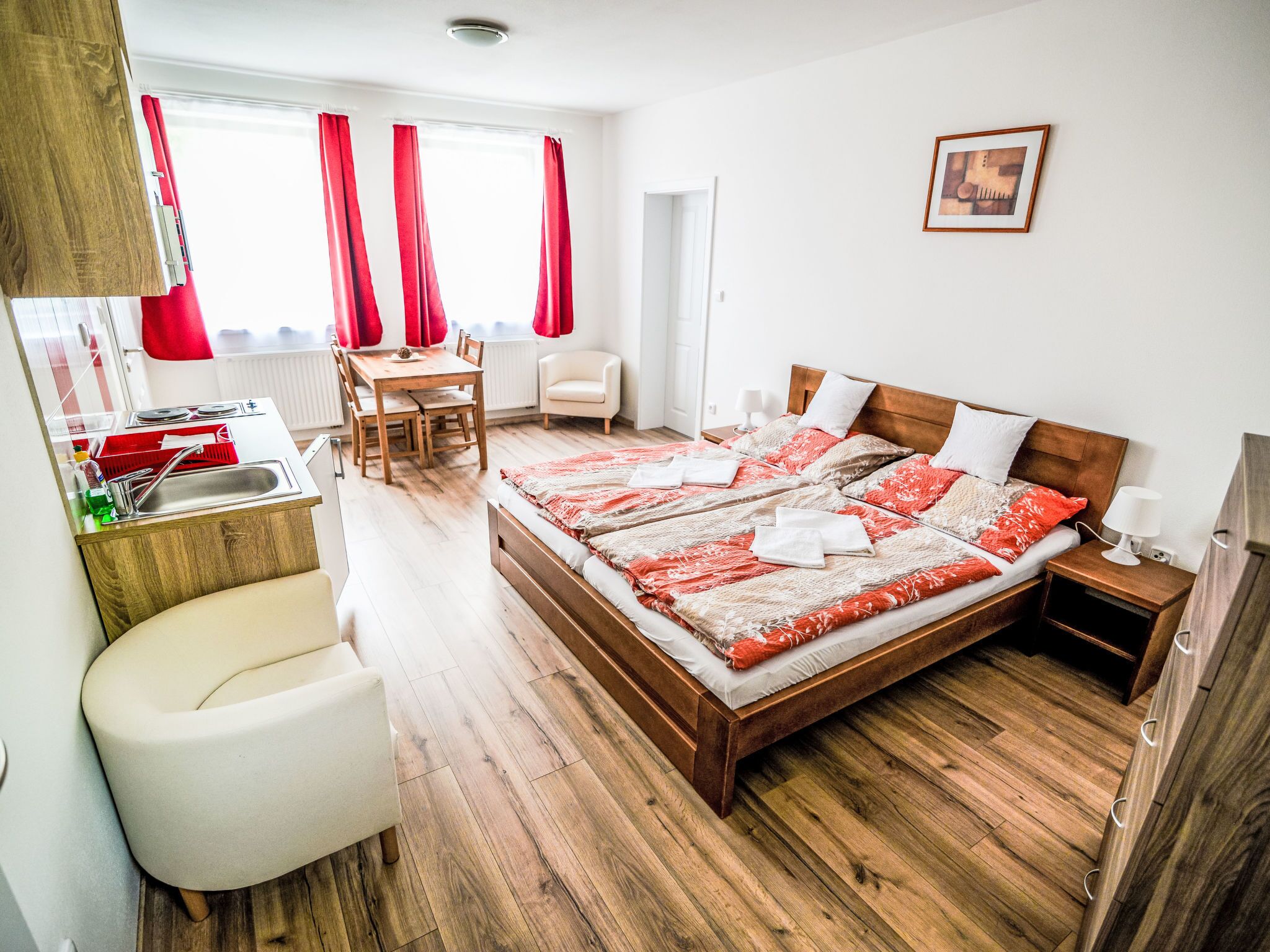 A hotel room with a wooden floor, a bed with red and white bedding, a small desk with a chair, a white armchair, and a picture on the wall.