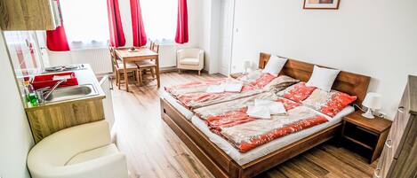 A hotel room with a wooden floor, a bed with red and white bedding, a small desk with a chair, a white armchair, and a picture on the wall.