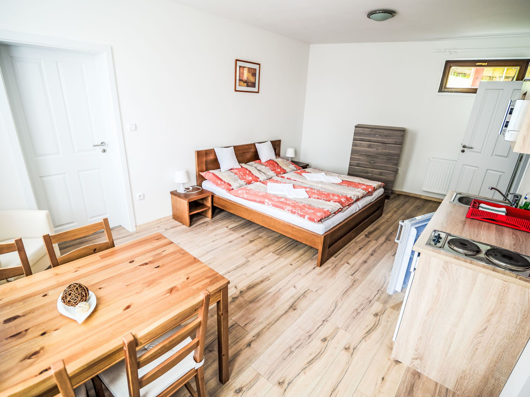 Un dormitorio con una cama de madera, mesitas de noche, una lámpara pequeña de noche, una mesa de comedor de madera con sillas, y una zona de cocina con un fregadero y una estufa.