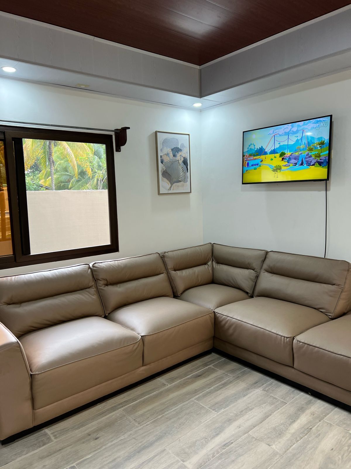 A corner living room with a beige sectional sofa, a framed abstract painting, and a colorful landscape TV on the wall.