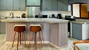 A modern kitchen with a wooden ceiling, grey cabinets, a central island with stools, and a stainless steel refrigerator.