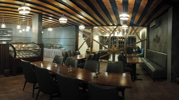 A restaurant with wooden tables and chairs, a dessert display case, and hanging pendant lights. - Lagoon Lodge (Hithadhoo)