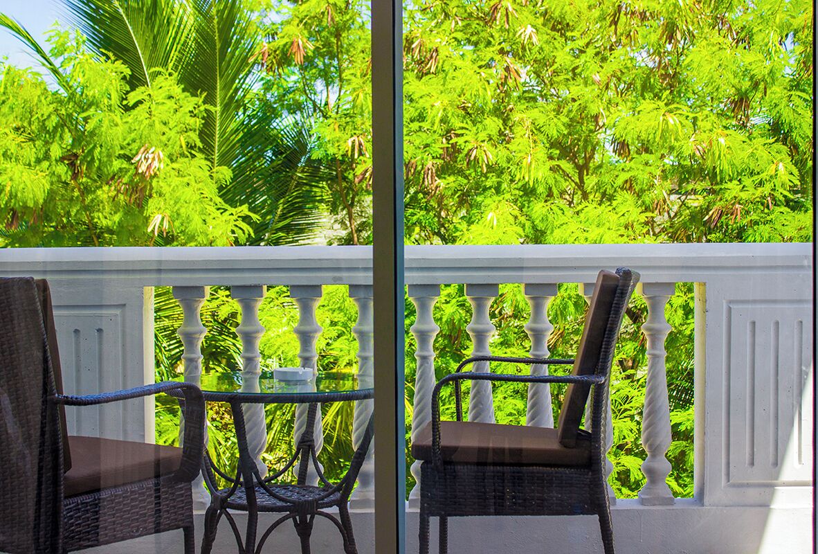 Un balcon agrémenté de deux fauteuils en osier et d’une table en verre, le tout entouré d’un luxuriant feuillage verdoyant.