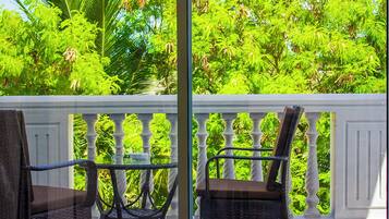 Un balcon agrémenté de deux fauteuils en osier et d’une table en verre, le tout entouré d’un luxuriant feuillage verdoyant.
