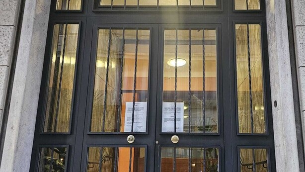 A building entrance with a large, dark-framed window with vertical bars, a stone wall, and a visible sign inside.
