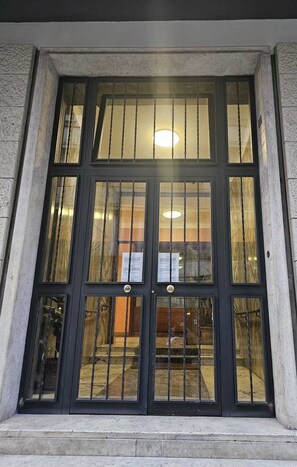 A building entrance with a large, dark-framed window with vertical bars, a stone wall, and a visible sign inside.