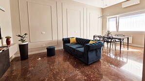A spacious living room with a dark blue sofa, a dining table with chairs, and a large window providing natural light.