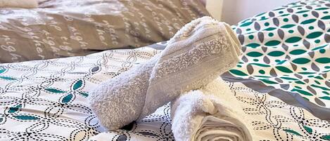 A neatly made bed with a patterned bedspread, a rolled-up towel, and a pillow with a text pattern.