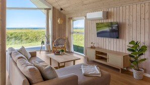 A spacious living room with a large window, wooden walls, a sofa, a TV, and a plant. - "Tilje" - 80m from the sea by Interhome (Hjørring)