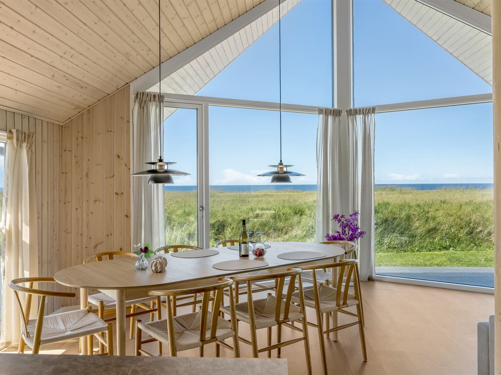 Un espace repas avec une grande table et des chaises, de grandes fenêtres donnant sur la mer et des luminaires suspendus.