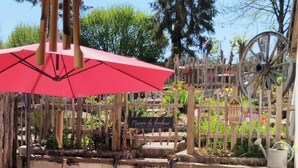 Un área de estar al aire libre de estilo rústico con una mesa y bancos de madera, un paraguas rojo y una valla de madera con un letrero que dice "Maison du Bonheur".