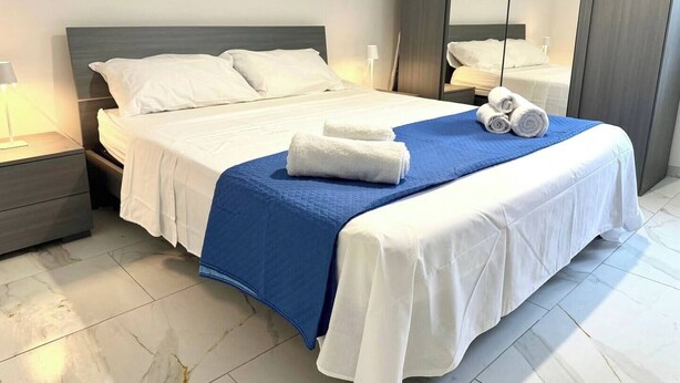 A neatly made bed with white linens and a blue accent, a nightstand with a lamp, and a large mirror reflecting the room. - Betta's House, the vacation home just a few kilometers from Taormina and Catania. (Sant'Agata li Battiati)