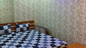 A bedroom with a bed, wooden headboard, patterned wallpaper, and a nightstand. - Dev In Guest House (Varanasi)