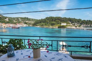 Un balcon avec une table dressée pour le dîner, offrant une vue sur une ville côtière aux eaux d’un bleu limpide, avec des collines en arrière-plan.