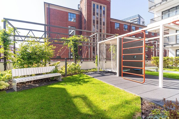 A playground with a climbing structure, a bench, and a grassy area in front of a building with brick walls and windows.