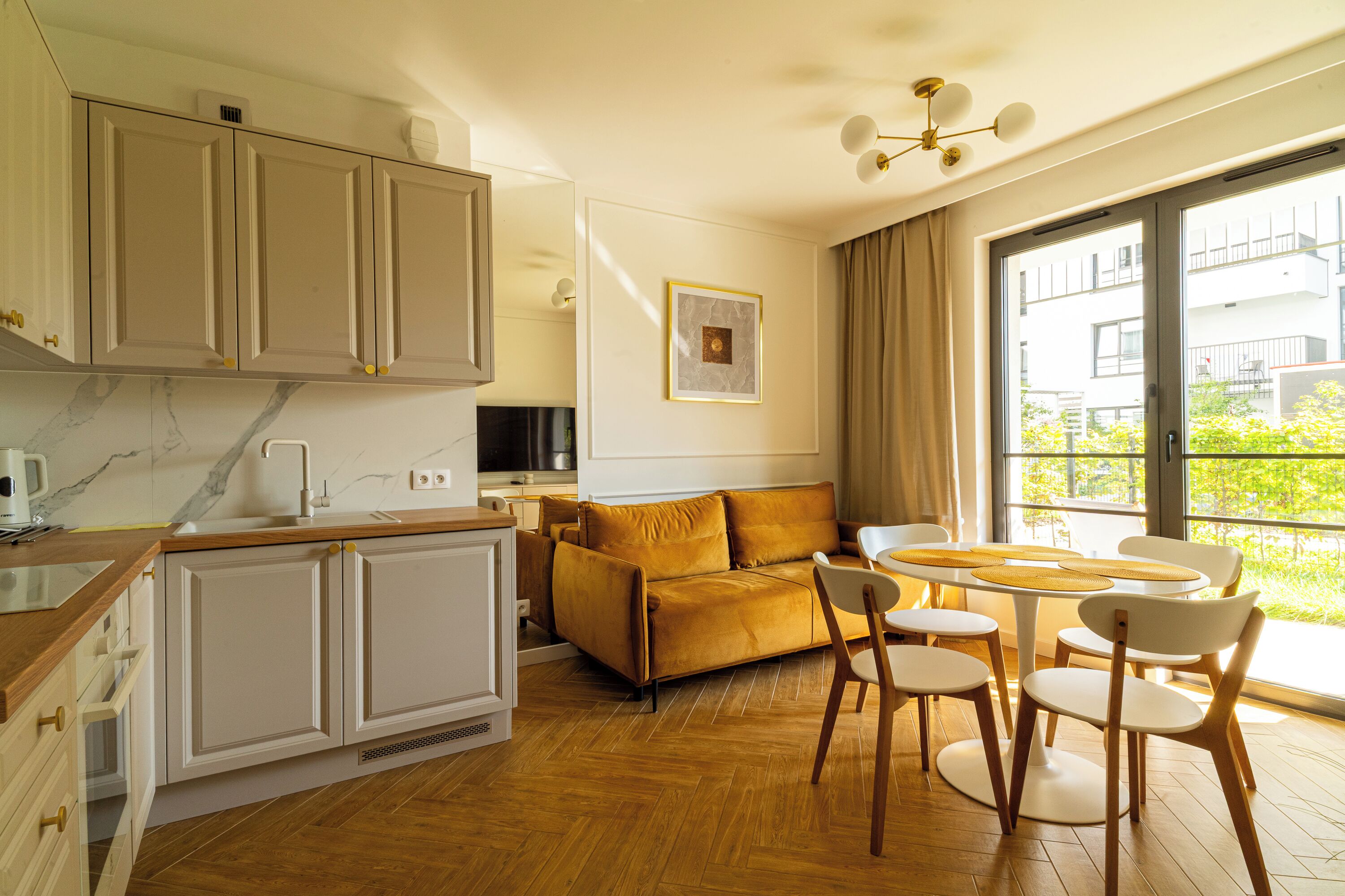 Eine moderne Küche mit weißen Schränken, einer Holz Arbeitsplatte und einem eingebauten Herd. Im Wohnbereich befindet sich eine Couch und Stühle, sowie ein großes Fenster mit Blick auf Grünflächen.