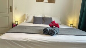 A neatly made bed with white linens, a grey blanket, and rolled grey towels. There are two bedside lamps with patterned shades, a wall-mounted decorative item, and a green curtain. - Apartment Tahiti near hospital, wifi,A&C,Swimming pool (Pirae)