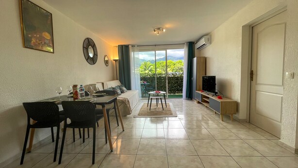 A living room with a dining area, a TV, and a sliding glass door leading to a balcony. - Apartment Tahiti near hospital, wifi,A&C,Swimming pool (Pirae)