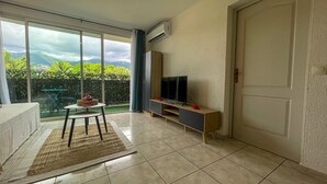 A modern living room with a TV, a small table, and a view of greenery through large windows. - Apartment Tahiti near hospital, wifi,A&C,Swimming pool (Pirae)