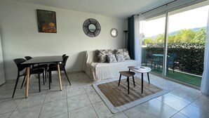 A modern living room with a dining area, a sofa with patterned cushions, a coffee table, and a large sliding glass door leading to a balcony with a view of greenery and hills. - Apartment Tahiti near hospital, wifi,A&C,Swimming pool (Pirae)