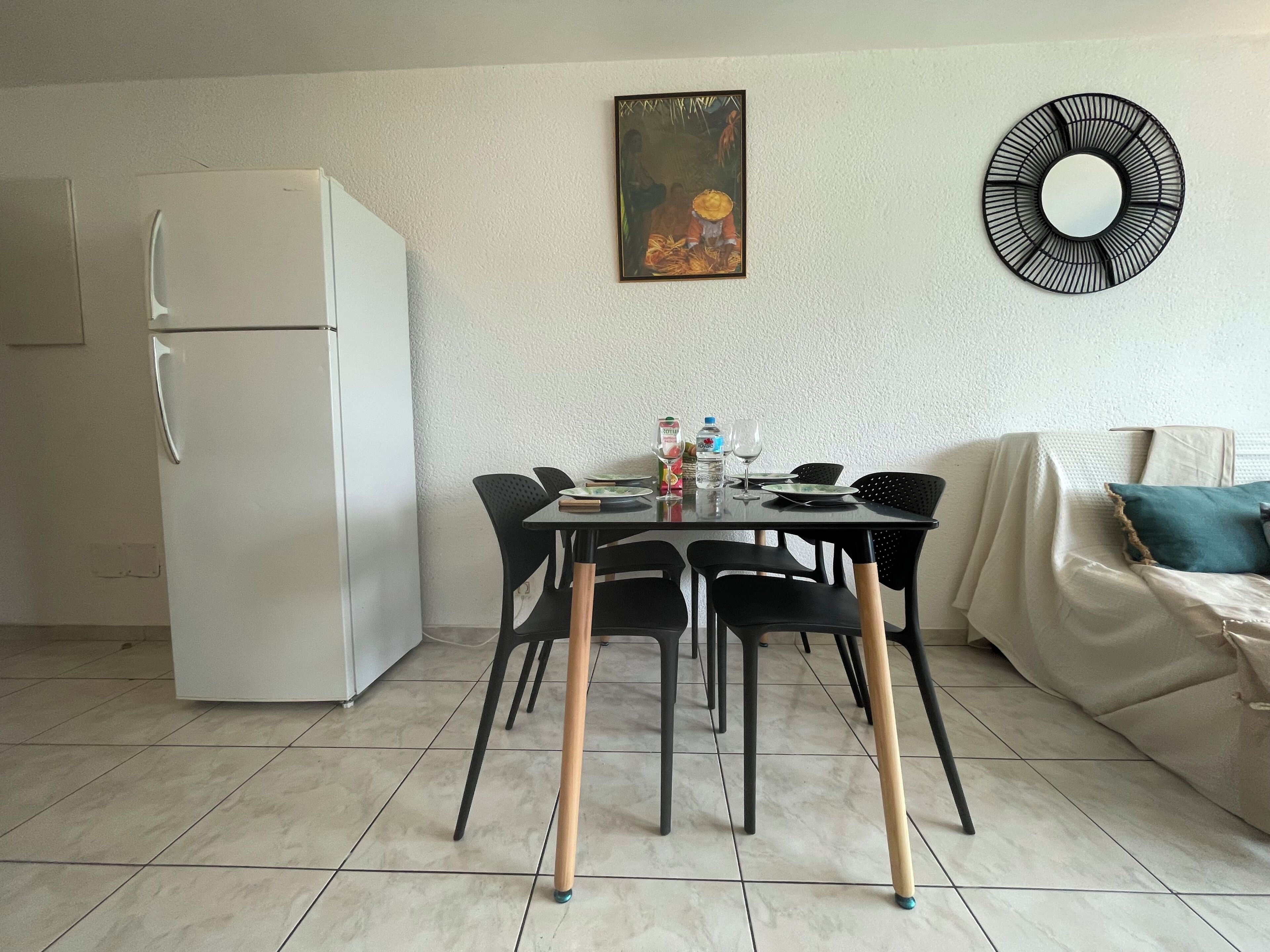 Cocina compacta con un refrigerador blanco, una mesa de comedor con sillas negras y un sofá con un cojín verde azulado.