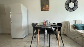 Cocina compacta con un refrigerador blanco, una mesa de comedor con sillas negras y un sofá con un cojín verde azulado.