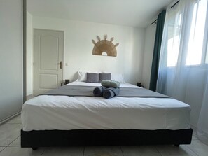 Uma cama bem arrumada com lençóis brancos e toalhas enroladas, um objeto decorativo na parede, uma porta e uma janela com cortinas.