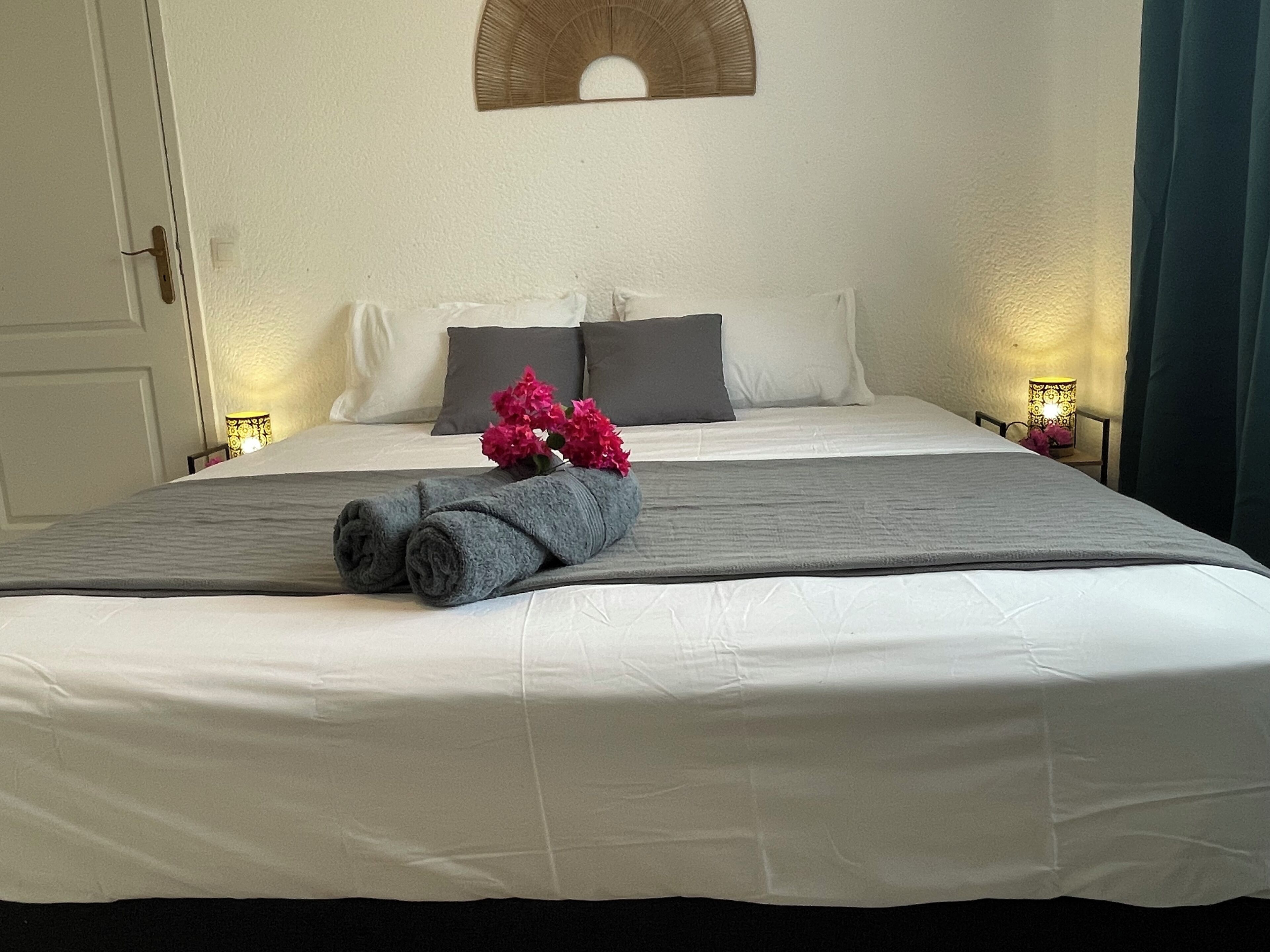 A neatly made bed with a rolled towel and flowers, a bedside lamp, and a wall-mounted mirror.