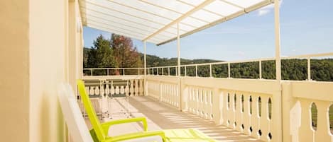 A covered outdoor seating area with white and yellow chairs, a railing, and a view of a forested hillside.