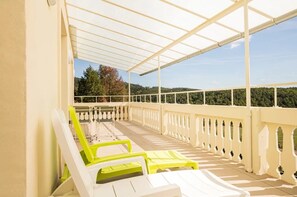 A covered outdoor seating area with white and yellow chairs, a railing, and a view of a forested hillside.