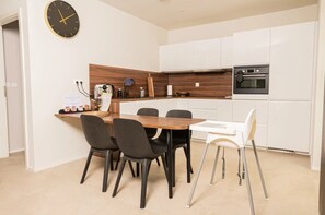 A modern kitchen with white cabinets, a wooden countertop, and a black clock. There is a dining table with four chairs, one of which is white.