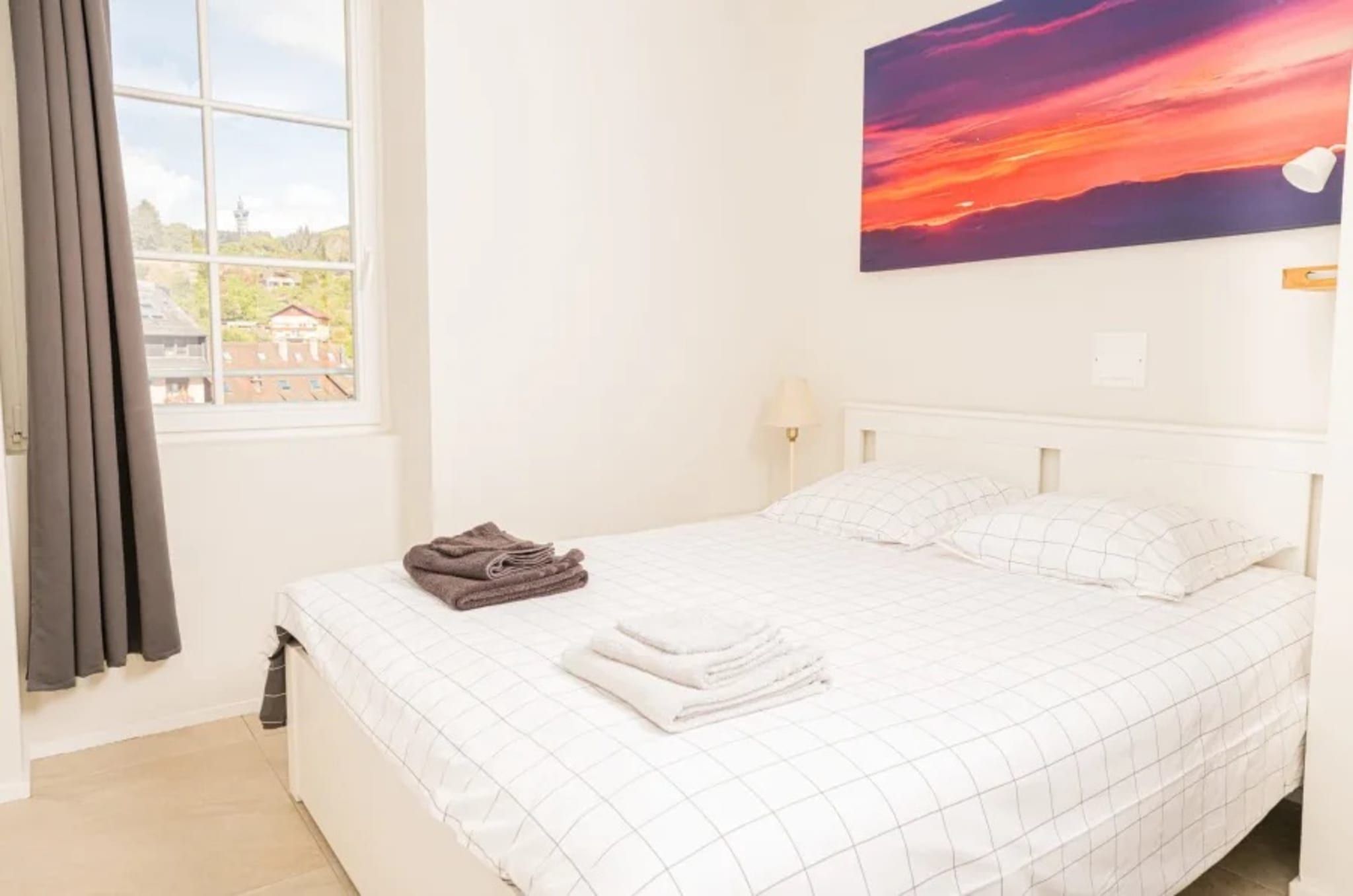 Ett hotellrum med en säng, ett fönster med gardiner, en tavla på väggen och utsikt över byggnader utanför.