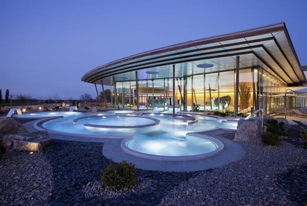 Relax in a serene atmosphere surrounded by natural elements and open spaces, featuring a curved glass facade and multiple hot tubs.
