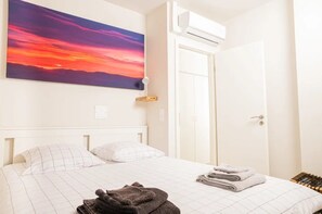 A bedroom with a bed, a wall-mounted air conditioner, a door, and a painting of a sunset.