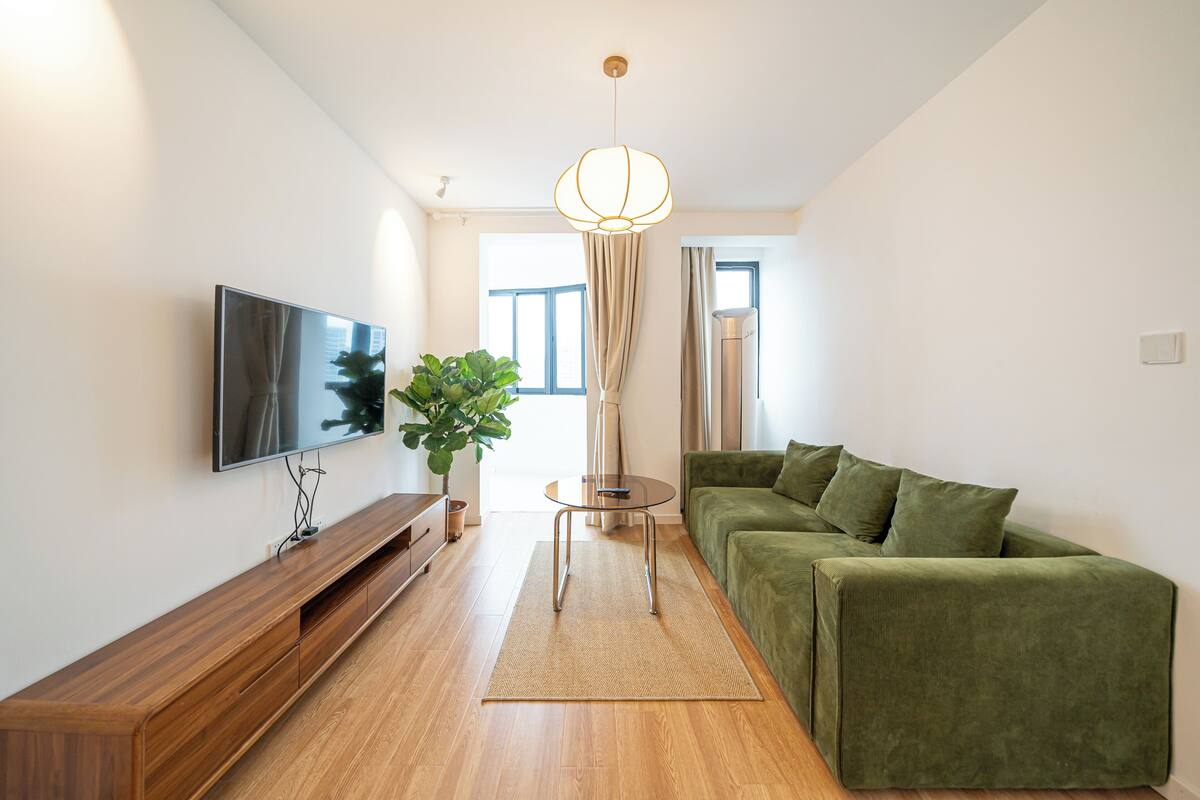 A modern living room with a flat-screen TV, a green sofa, a wooden TV stand, a small side table, and a large window with curtains.