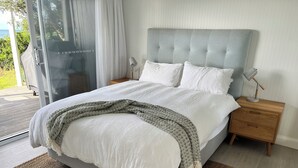 A bedroom with a large bed, white bedding, a grey headboard, two bedside tables with lamps, and a view of the outdoors through a glass door.