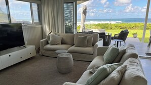 A modern living room with a large TV, beige sofas, a coffee table, and a view of the ocean.