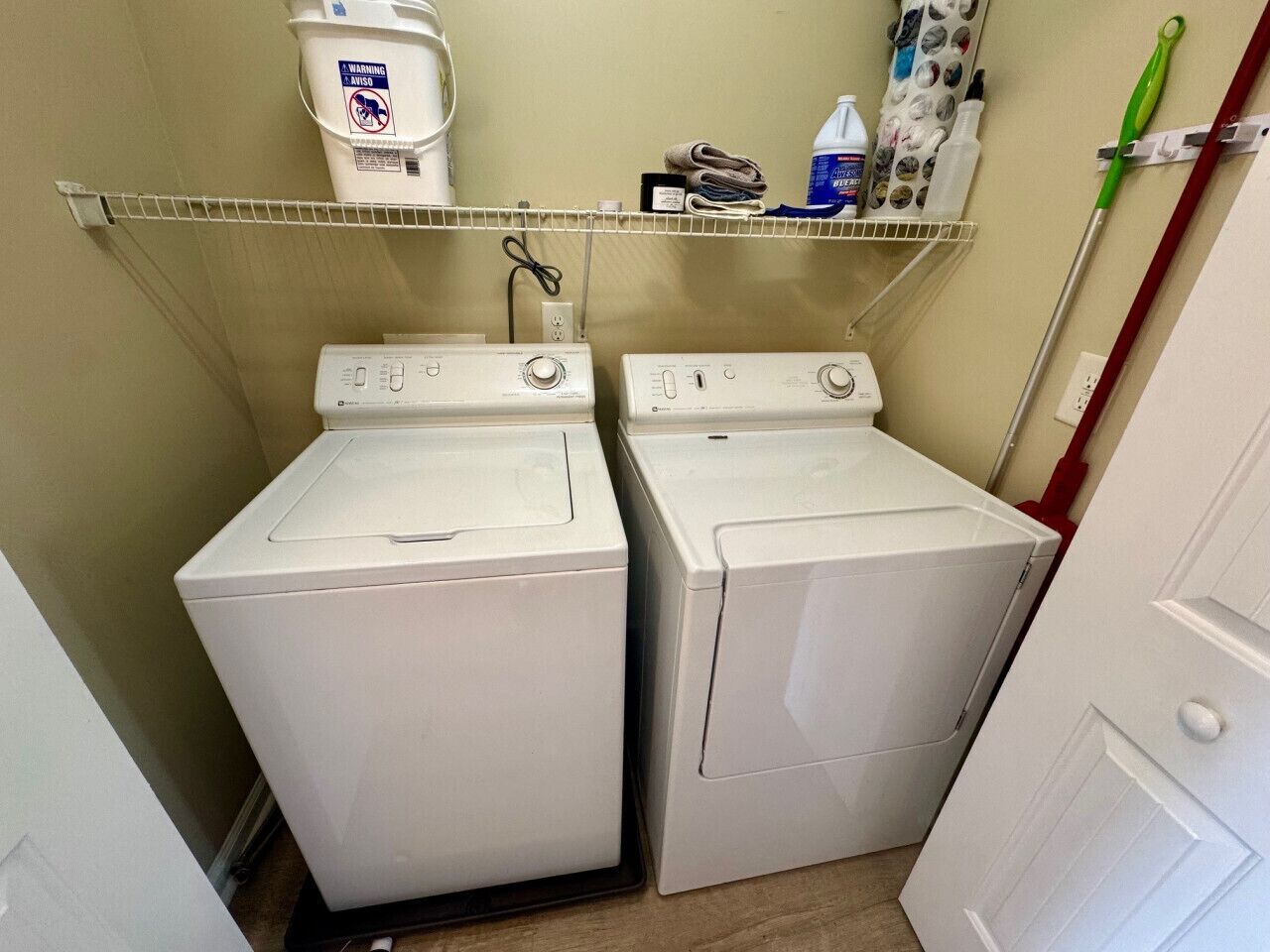 Un espace buanderie comprenant deux lave-linge et deux sèche-linge à chargement frontal, tous deux de couleur blanche, une étagère avec des produits de nettoyage et une serpillière accrochée au mur.