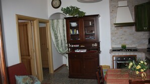 A compact living space with a dining area, a kitchen with a stove and cabinets, a wooden cabinet with glass doors, a clock on the wall, and a plant on the cabinet. - Carrube House, in Andrano offers a terrace and sea views. (Marina di Andrano)