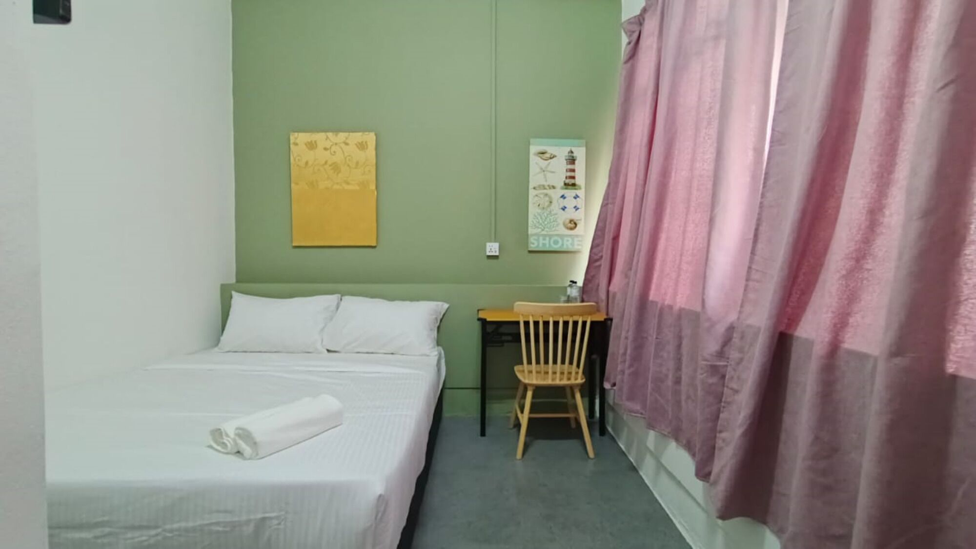 Una cama individual con una toalla blanca enrollada, una silla de madera, un escritorio pequeño y una pared con un letrero que dice 'SHORE'.