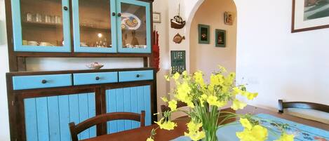 A dining area with a wooden table, chairs, and a blue tablecloth. There is a vase with yellow flowers on the table.