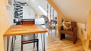 A cozy loft bedroom with a wooden dining table, a brown leather chair, and a view of a kitchen and living area. - La Maison Bleue, on the Bay of the Somme, in a peaceful natural setting (Pendé)