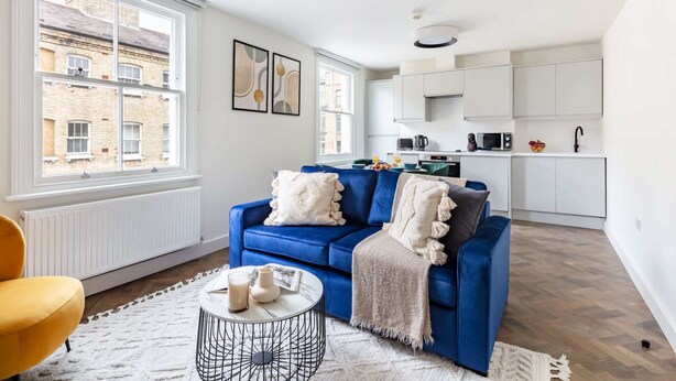 A modern living room with a blue sofa, a yellow armchair, and a white coffee table. A kitchen with white cabinets and appliances is visible in the background. - Bright 1BR Near Museum of London (London)