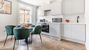 A modern kitchen with white cabinets, a dining table with chairs, and a window with a view of buildings. - Bright 1BR Near Museum of London (London)