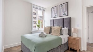 A bedroom with a bed, a bedside table with a lamp, and a window with a view of buildings. - Bright 1BR Near Museum of London (London)