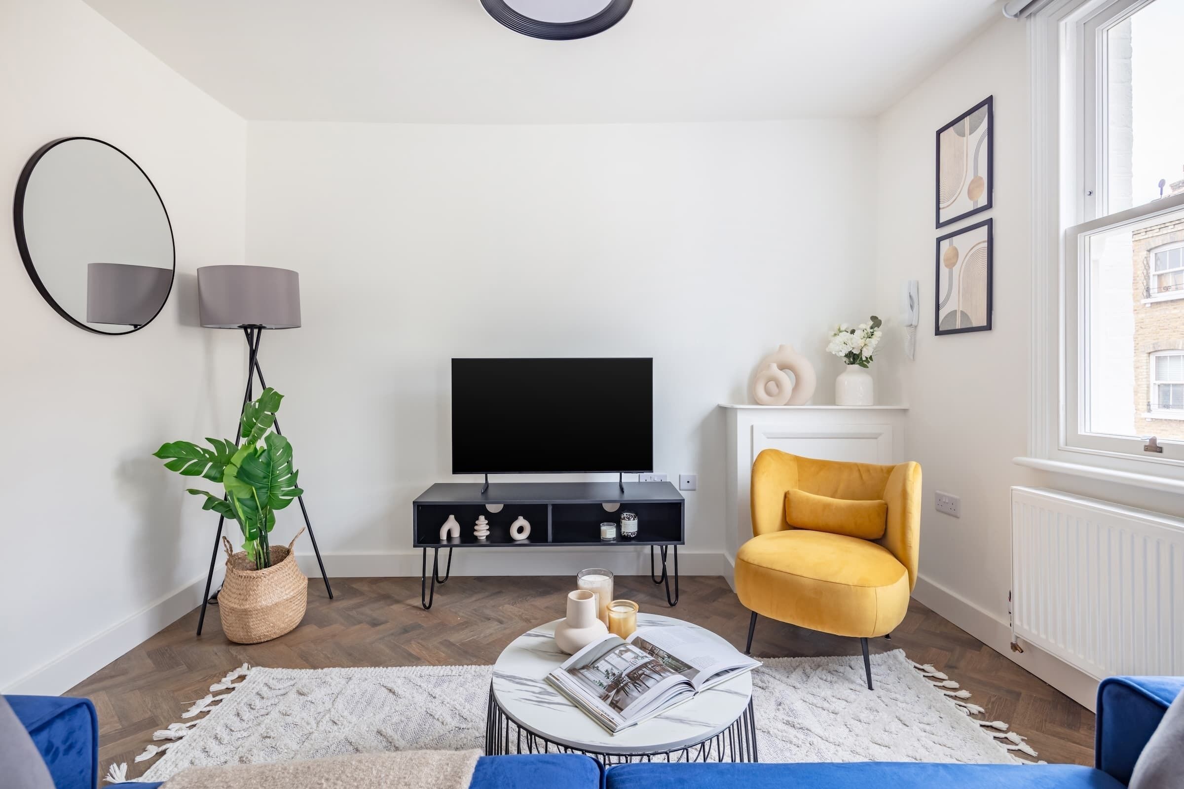 A modern living room with a flat-screen TV, a yellow armchair, a coffee table with magazines, a potted plant, and a large mirror.