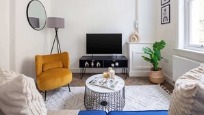 Un salón con un sillón amarillo, una mesa de centro redonda con revistas, un mueble de televisión negro, una planta en maceta y una ventana con vistas.