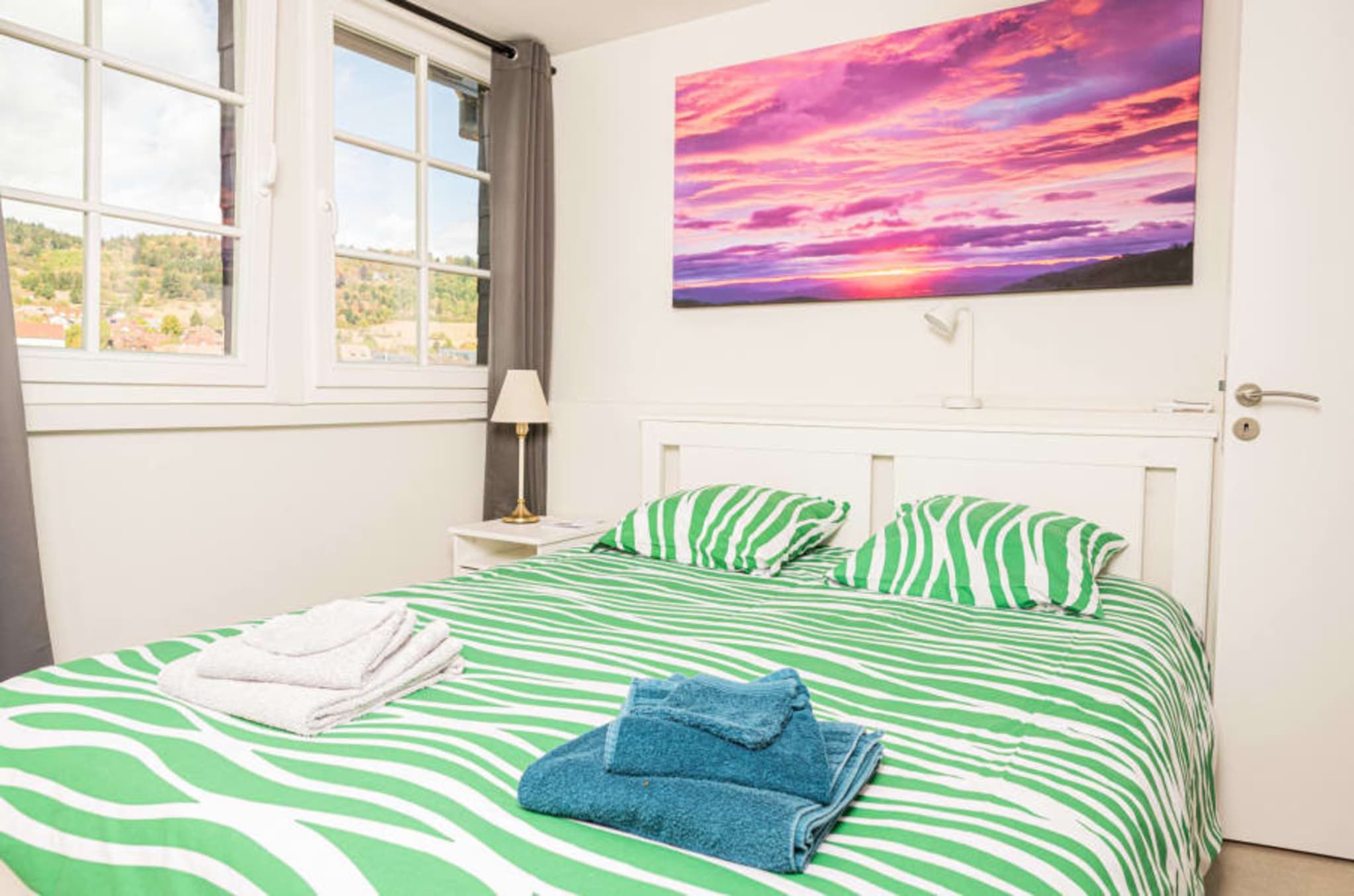 A bed with green and white striped bedding, a white bedside table with a lamp, and a painting of a sunset on the wall.