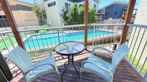 Une terrasse en bois avec une table ronde et deux chaises blanches, donnant sur une piscine et des immeubles d’habitation.