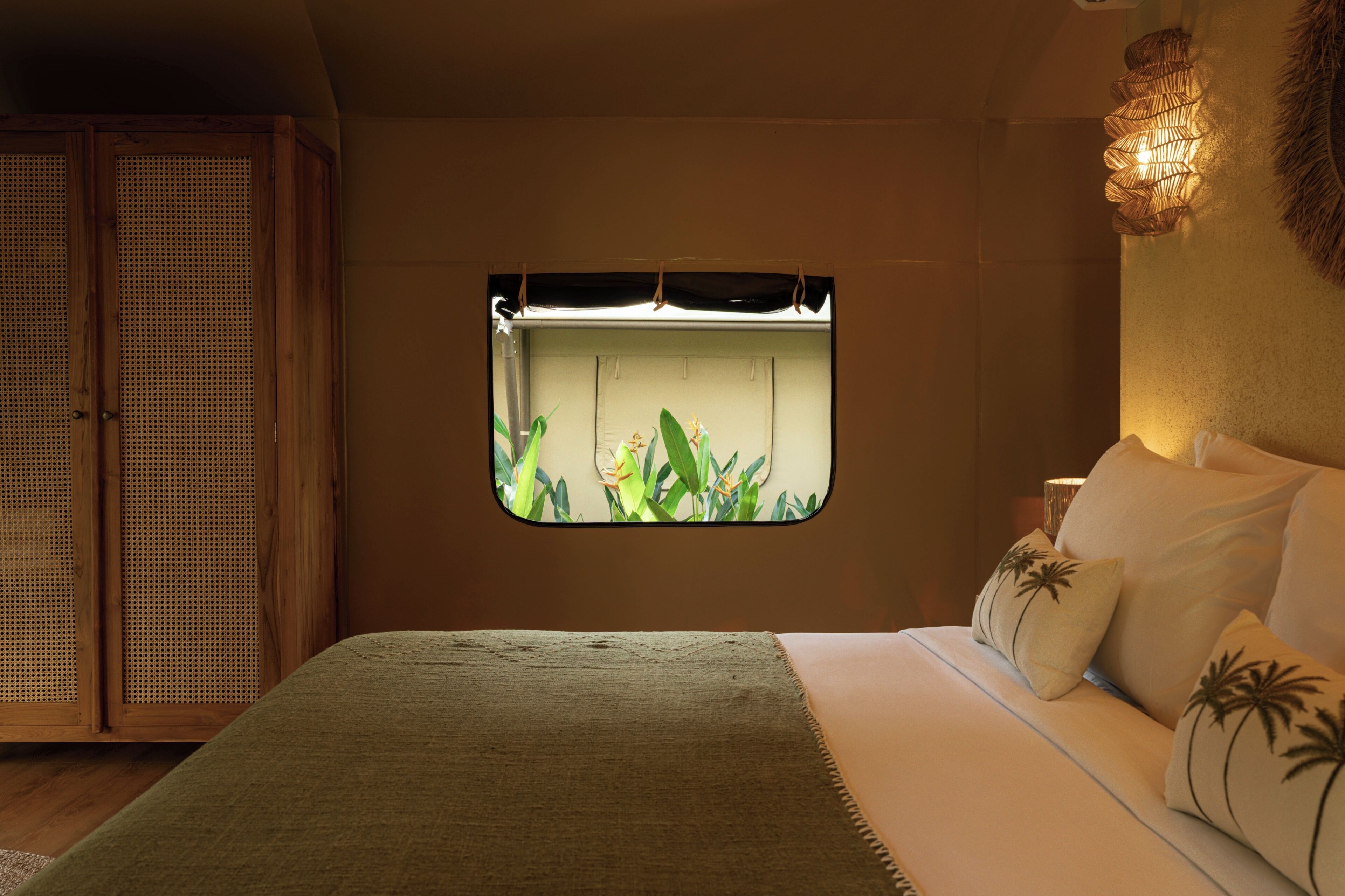 A bedroom with a bed, a window with a view of plants, a wooden wardrobe, and a wall-mounted light fixture.